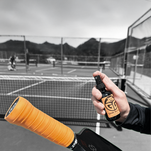 pickleball court background with a hand spraying the grizzlee gripz overgrip with enhanced grip spray